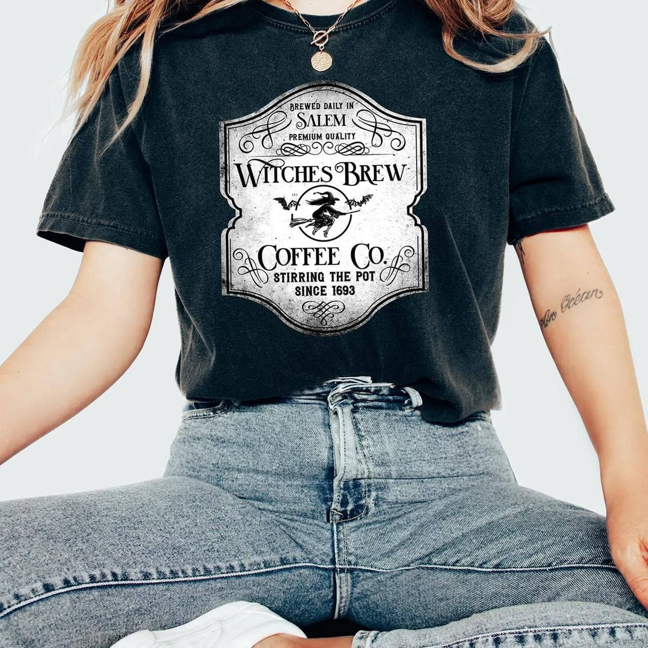 A woman wearing a black t-shirt with a vintage "Witches Brew Coffee Co." logo depicting a witch on a broom.